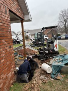 Two workers are in a trench near a brick house foundation, with construction equipment and vehicles parked nearby on a cloudy day.