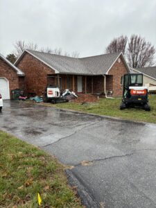 A brick house with construction equipment and materials on the front lawn, including two Bobcat machines, on a cloudy, wet day.