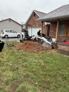 A small excavator digs near the front porch of a brick house, with a pile of dirt and tools scattered on the lawn. A white car is parked nearby.