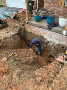 A person is digging and working with pipes in a deep trench next to a brick house and porch, surrounded by soil, tools, and potted plants.
