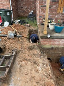 A person is working in a deep trench next to the foundation of a brick house, surrounded by soil, tools, and construction equipment.