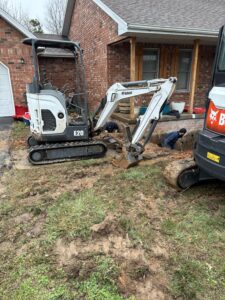 A small excavator is digging in front of a brick house, with workers nearby and disturbed soil and grass in the yard.