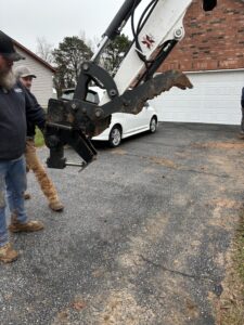 Two men stand near a mechanical excavator claw on a driveway in front of a white car and a garage.
