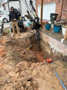 A construction worker stands by a mini excavator digging a large hole beside a brick house and porch, with tools and planters visible nearby.