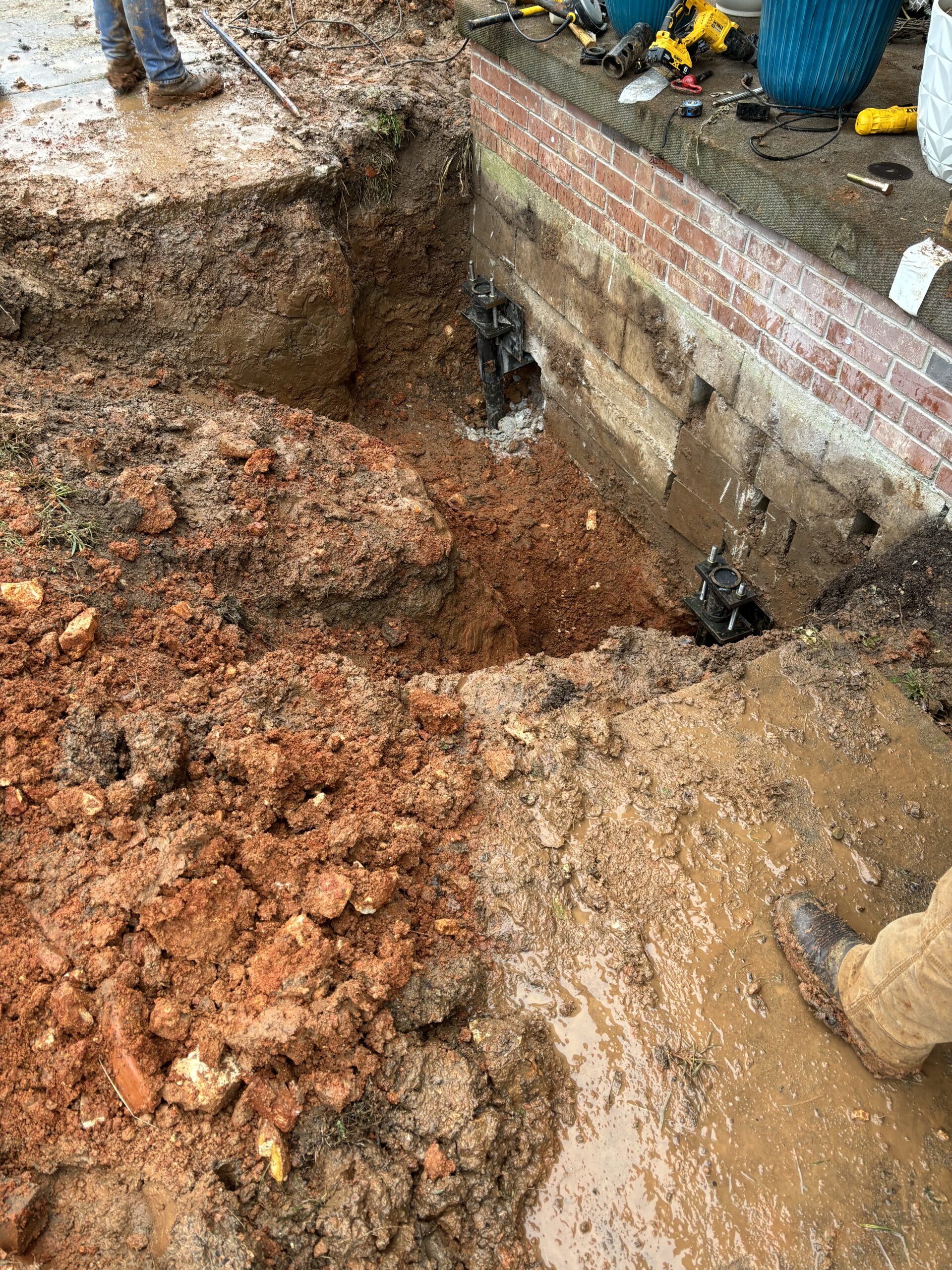 Exposed foundation of a brick building with large dug-out sections of soil and visible support piers during repair work; tools and equipment are nearby.