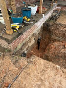 A large hole is dug next to a brick house foundation, with tools and planters on the porch above, showing ongoing foundation repair work.