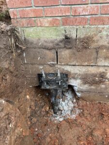 A steel foundation pier is installed beneath a cracked brick wall, with exposed soil and concrete visible around the base of the building.