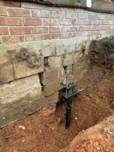 A house foundation with exposed soil shows a section of the wall and an installed steel pier used for structural support.