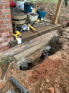 A section of a brick porch with tools, flower pots, and a dug-out area exposing the foundation and support jacks beneath the structure.