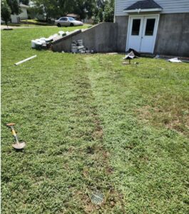 A patch of grass with a visible trench line leads to a basement entrance; tools and construction materials are scattered nearby.