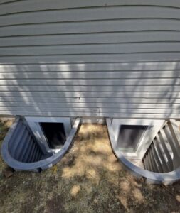 Two basement window wells with metal liners are set in the foundation of a house with light-colored vinyl siding. The ground around the wells is dirt and sparse grass.