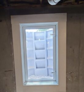 A small basement window with a white frame looks out onto a gray window well lined with gravel at the bottom.