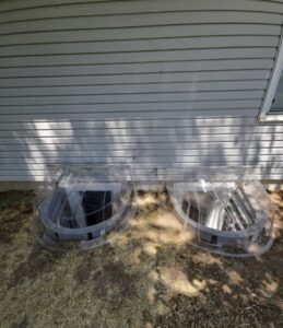 Two clear plastic window well covers placed over basement window wells beside a gray-sided house with a small window visible on the right.