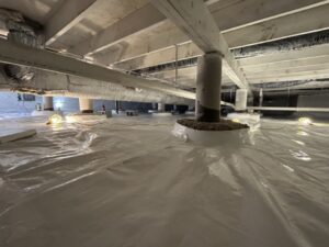 Crawl space with white vapor barrier installed on the floor and pillars, exposed beams above, and several work lights illuminating the area.