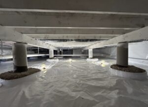 View of a crawl space with concrete support pillars, wooden beams, and a plastic vapor barrier covering the floor; LED work lights illuminate the area.