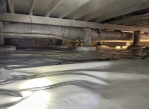A crawl space with a plastic vapor barrier on the ground, exposed pipes, and beams overhead, illuminated by a flashlight.