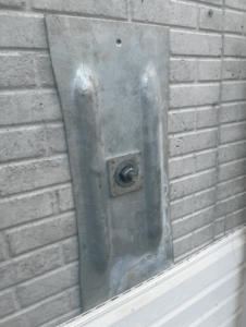 A corrugated metal plate with a central bolt is secured to a brick wall above white siding.