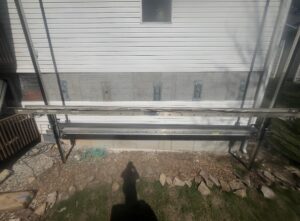 A construction scaffold is set up against the side of a white house with mounting brackets attached to the wall and a green garden hose on the ground below.