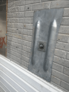 A metal plate with a protruding bolt is attached to a gray brick wall above white siding.