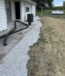 A gravel path is laid along the side of a white house, leading from a concrete porch to a yard area with straw and grass. A black hose runs near the house.