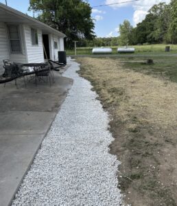 White gravel borders the side of a white house with a concrete patio and outdoor furniture, leading to a yard with grass, dirt, and two propane tanks in the background.