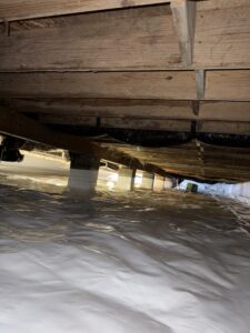 View of a crawl space with a white vapor barrier on the ground, wooden beams above, and support columns spaced throughout the area.
