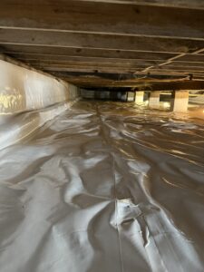 A crawl space with a plastic vapor barrier covering the floor and partially up the walls, wooden beams overhead, and some support columns visible.