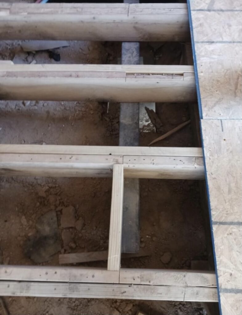 Wooden floor joists under construction with exposed beams and plywood partially covering the structure. Dirt ground is visible beneath the joists.