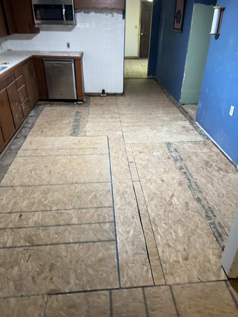 Kitchen with bare plywood subfloor, wooden cabinets, a wall-mounted microwave, and a dishwasher. Walls partially painted blue and white tiles near sink area.