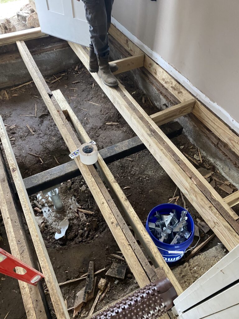 Wooden floor joists exposed during renovation with a person standing on them. A blue bucket with metal brackets and a roll of tape are visible on the site.