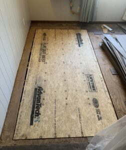 A partially renovated room shows new AdvanTech subfloor panels installed in one section, surrounded by exposed floor edges and stacked flooring materials nearby.