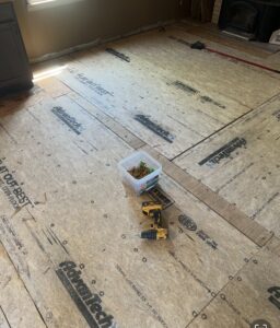 Plywood subfloor installation in progress with a plastic container of screws and a power drill placed on the floor.