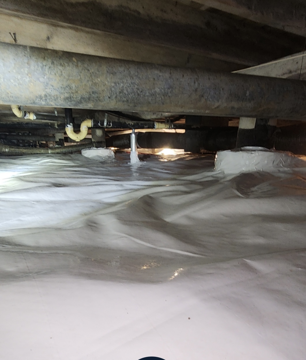 Plastic sheeting covers the floor of a crawl space with exposed pipes and beams visible overhead, illuminated by light in the background.