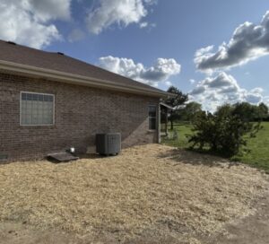 A brick house with a visible AC unit and fresh mulch covering the ground beside a newly completed crawlspace door installation, bordered by grass and shrubs under a partly cloudy sky.