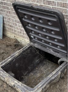 A large, open plastic underground utility box with a lid, located next to a brick wall—perfect for accessing services during crawlspace door installation—and partially surrounded by dirt.