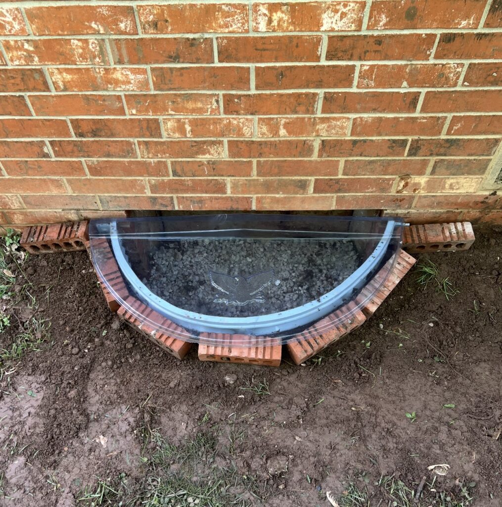 A window well with a clear plastic cover is installed against a brick wall, surrounded by loose bricks and soil.
