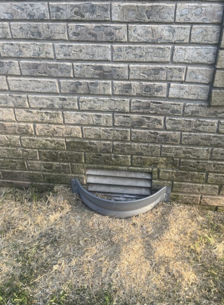 A broken or detached plastic window well cover lies on dry grass in front of a vent on a brick wall.