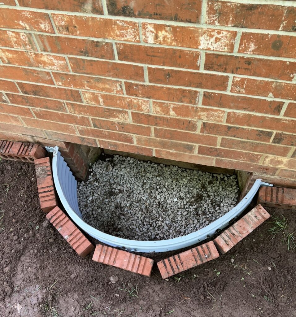 A metal window well ring installed against a brick house foundation, surrounded by loose gravel and bordered with bricks on the soil.