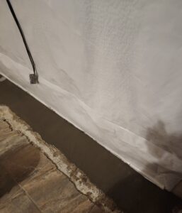 A basement wall covered with white plastic sheeting, a cable hanging near an outlet, and a strip of fresh concrete along the floor next to exposed tiling.