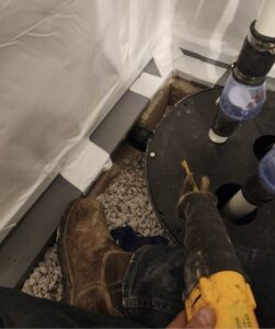Person using a reciprocating saw near a sump pump basin with exposed pipes, gravel, and a waterproof liner on the floor.