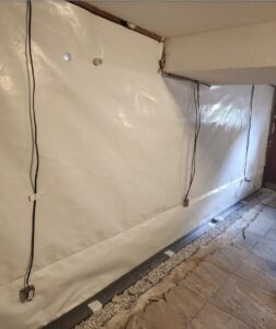A basement wall covered in white plastic sheeting, with electrical outlets, visible cables, and a gravel trench along the floor.