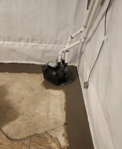 A sump pump system is installed in a basement corner with white pipes leading into the unit and fresh concrete surrounding the installation area.