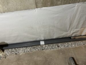 A basement wall with white waterproofing membrane, a black drainage channel at the base, and a layer of gravel along the concrete floor.