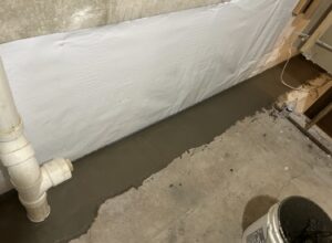 Basement floor with fresh concrete patch along the wall, white plastic sheeting on wall, exposed pipes, and a bucket in the foreground.