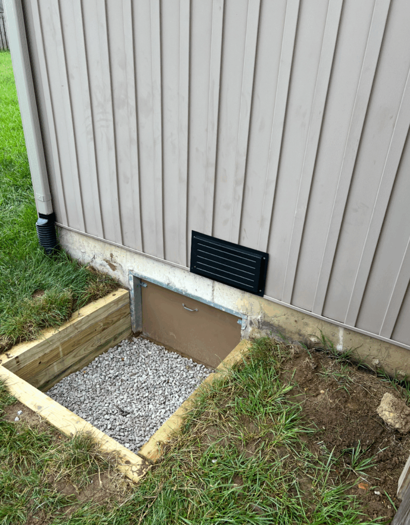 A ground-level crawl space entrance covered with a metal door, surrounded by wooden framing and gravel, next to a building with beige siding and a black wall vent above.