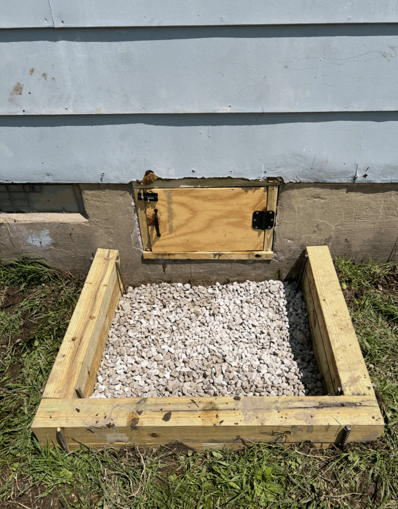 A small wooden frame filled with white gravel is positioned against the exterior wall of a house beneath a plywood access door.