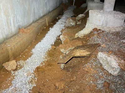 A narrow trench with loose gravel next to a concrete wall and uneven soil, scattered rocks, and construction debris.