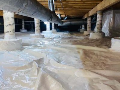 A crawl space with a plastic vapor barrier on the floor and support columns, featuring overhead ductwork and wooden beams.
