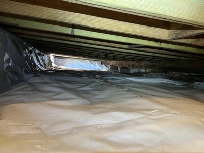View of a crawl space with a plastic covering on the floor, wooden beams above, and some light entering from an opening in the distance.