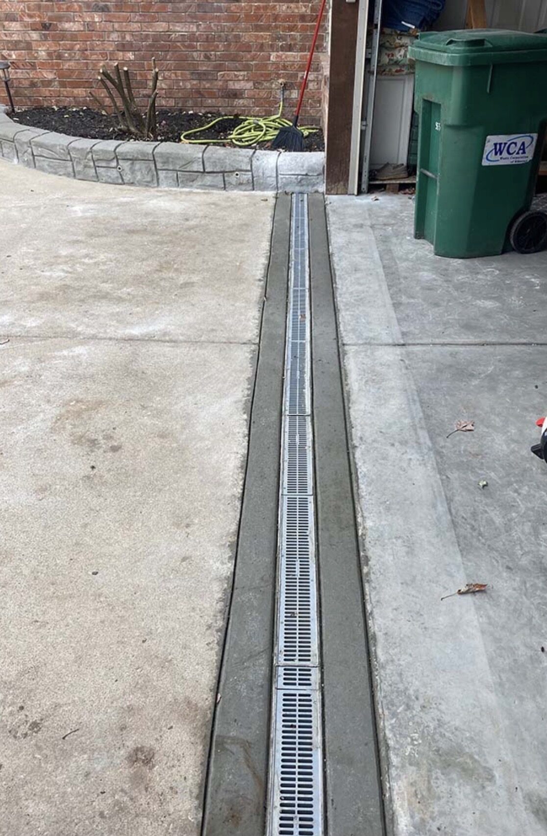 A metal drainage grate runs along the seam between concrete driveway and garage, with a green recycling bin and garden hose visible nearby.